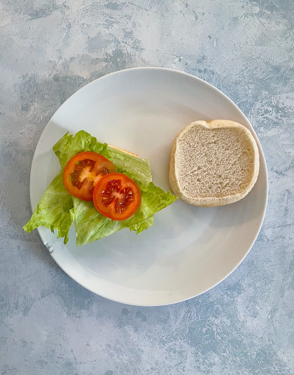burger bun on white plate, lid off, bottom topped with lettuce and 2 slices of tomato.