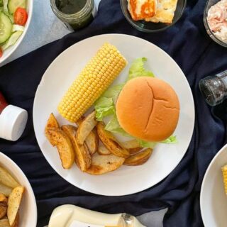 Burger on a plate with potato wegdes and corn on the cob. Ketchup, salt and pepper, coleslaw, salad and bowls of corn and wedges also on the table.