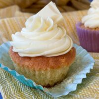 side view of a white chocolate cupcake topped with white chocolate buttercream and a square of white chocolate. Other cakes on glass stand in the background.
