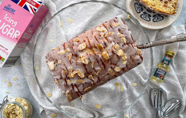 loaf cake topped with icing and toasted almonds on a glass cake stand with a silver slice. Slice of cake on plate to the side.