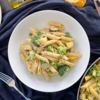 bowl of pasta with courgettes, peas and broccoli in a creamy sauce. frying pan to the side, garlic bread on plate, and jar of herbs also on table.