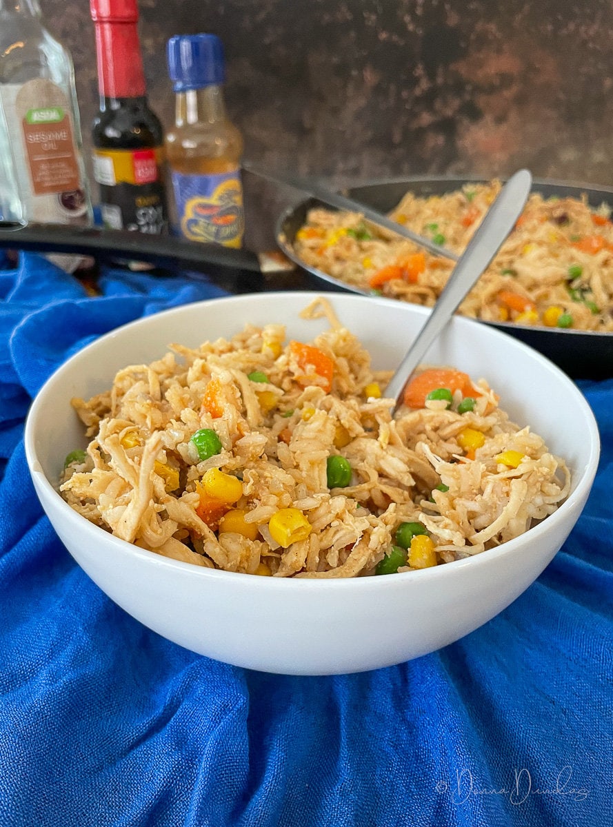 fried rice with chicken in small white bowl with fork. frying pan and bottles of sauces in the background.
