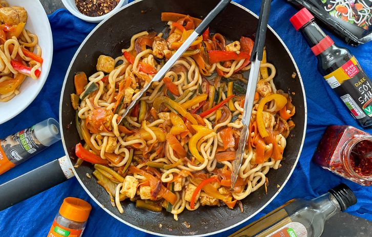 udon noodles, peppers, carrots and halloumi in a large frying pan with stir fry sauce. Soy sauce, chilli and other spices also on table.