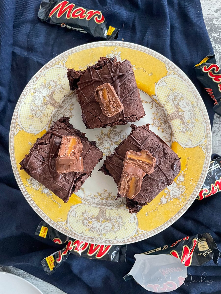 3 Squares of brownie topped with pieces of mars bar on a yellow patterned cake stand.