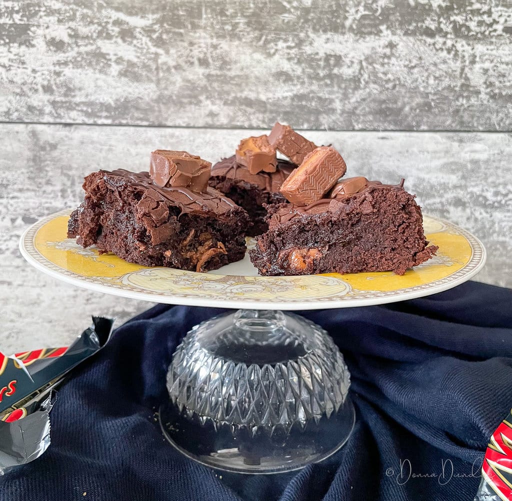 3 Squares of brownie topped with pieces of mars bar on a yellow patterned cake stand.