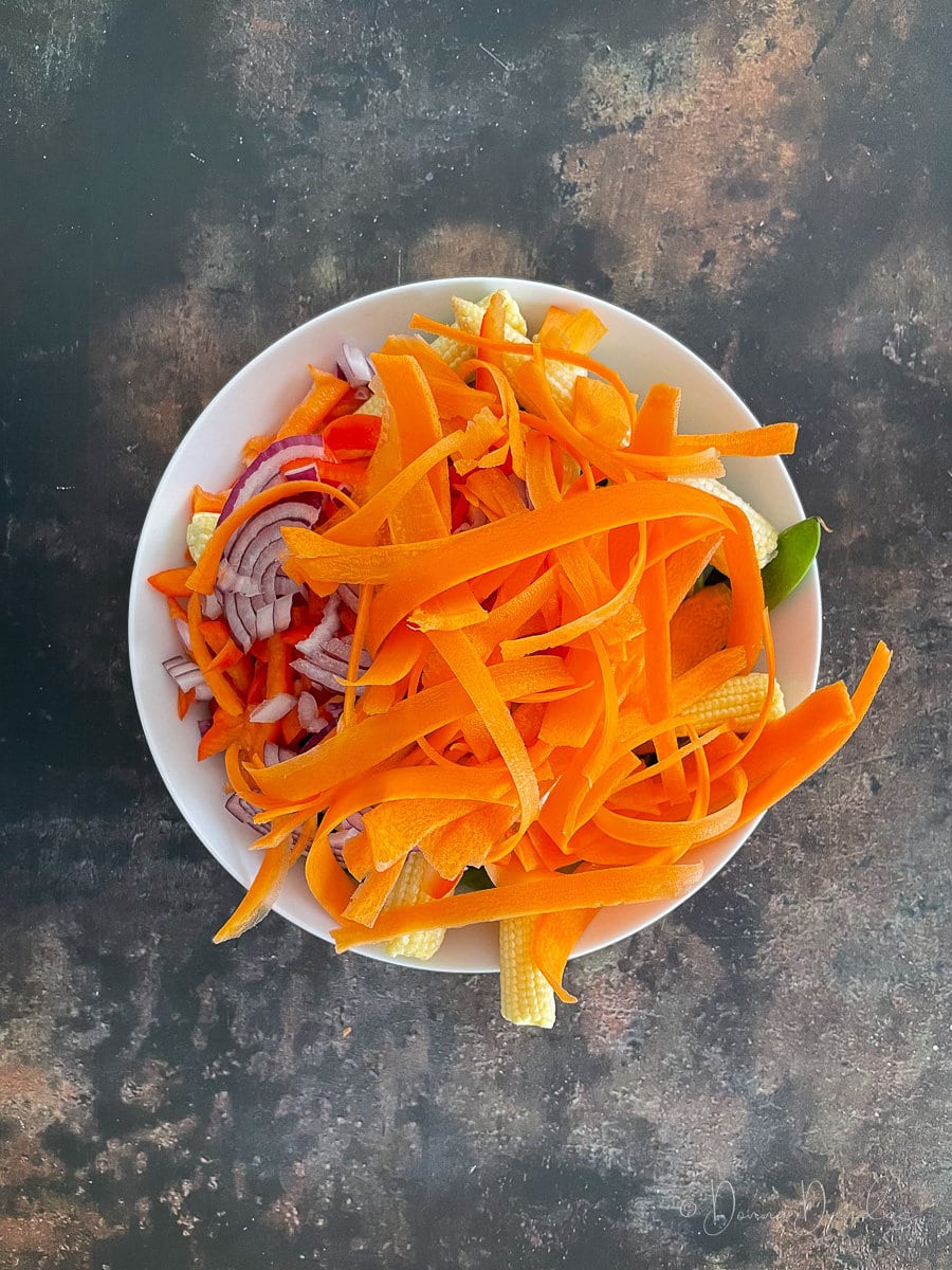 uncooked carrots, baby corn, onion, mangetout in white bowl.