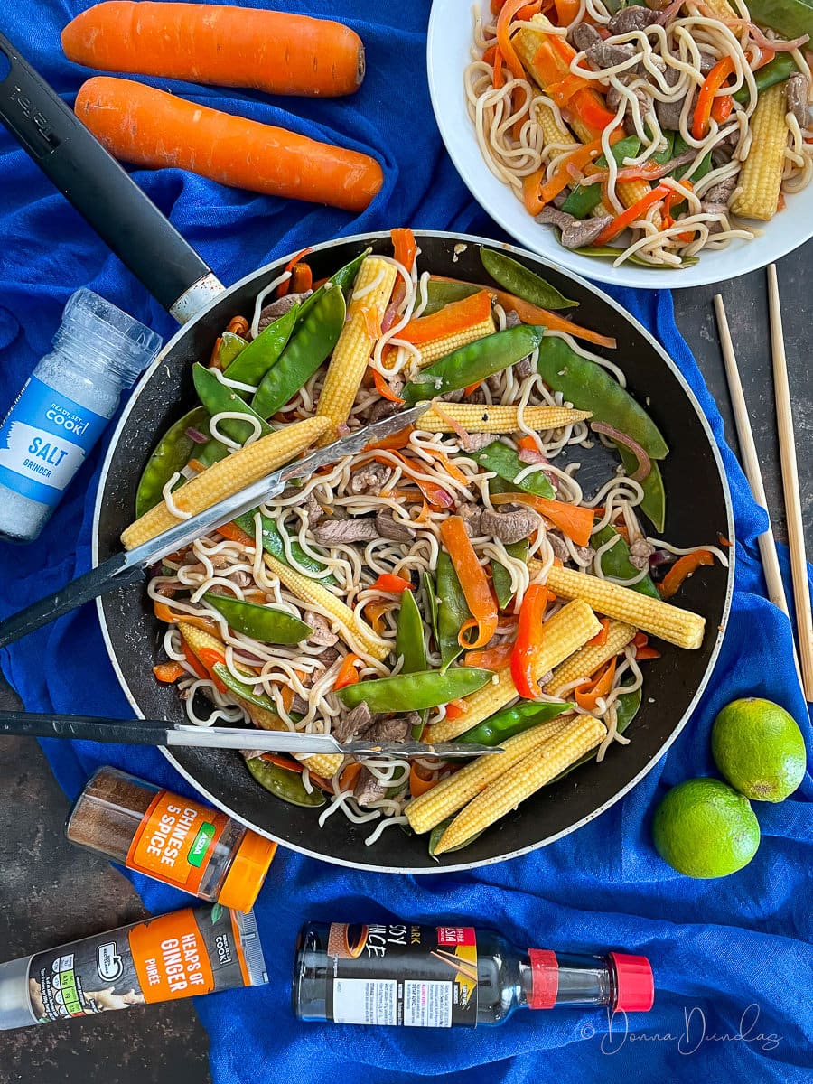frying pan with noodles,veg and lamb. Served portion, carrots, limes and soy also on table.