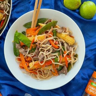 served portions of stir fry in white bowl with chopsticks. Frying pan, soy, 5 spuce and limes to the sides.