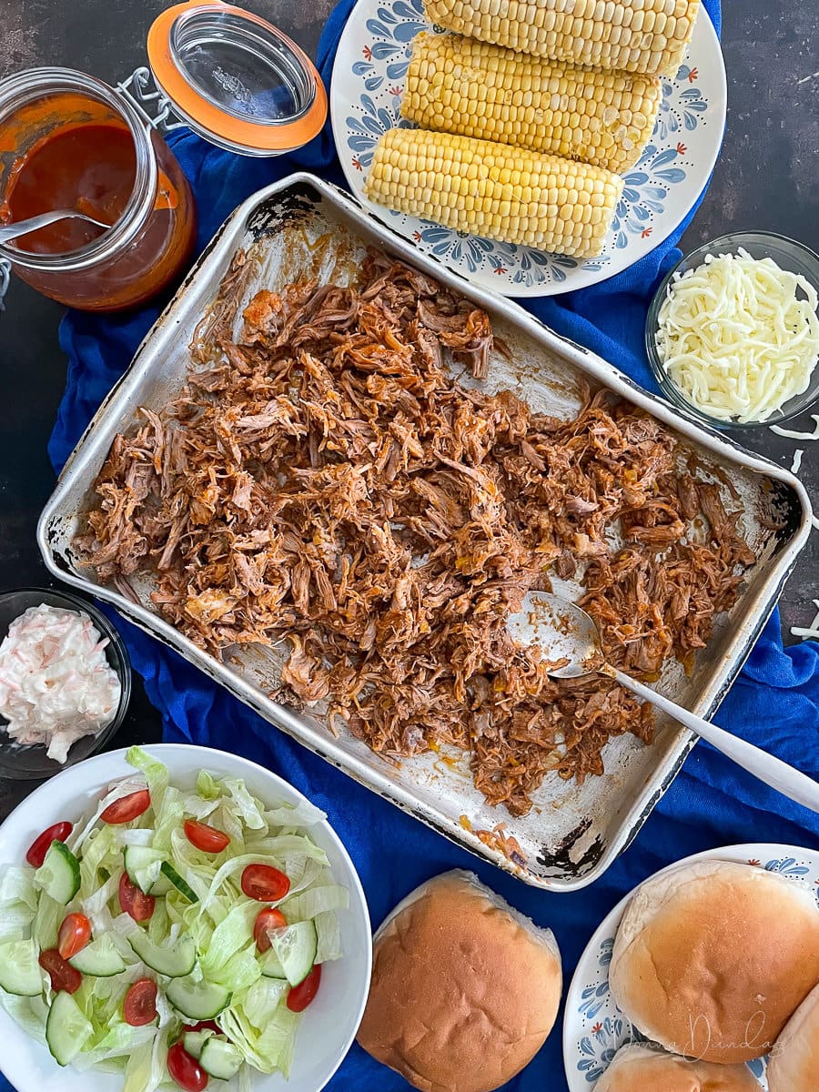 pulled pork on large silver baking tray with spoon. Corn, bread rolls, bbq sauce, salad and cheese also on table.
