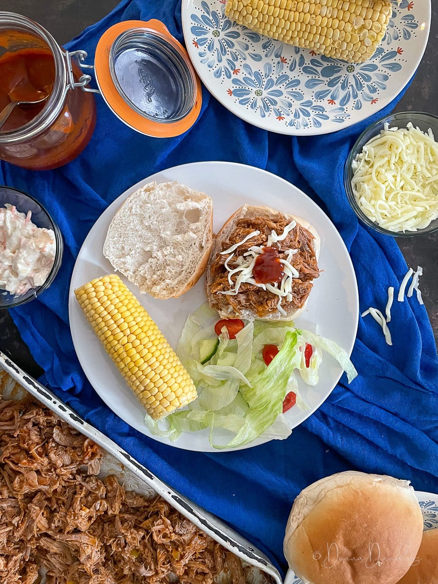 pulled pork on bread roll with grated cheese. Corn and salad also on plate, bbq sauce and tray of pork in background.