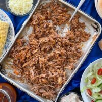 pulled pork on large silver baking tray with spoon. Corn, bread rolls, bbq sauce, salad and cheese also on table.