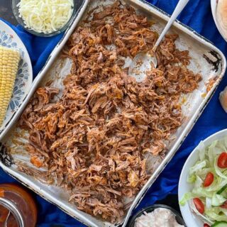 pulled pork on large silver baking tray with spoon. Corn, bread rolls, bbq sauce, salad and cheese also on table.
