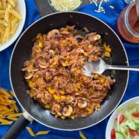 pulled pork pasta in frying pan with serving spoon. BBQ sauce, pasta bag, bowl of penne and salad also on table.