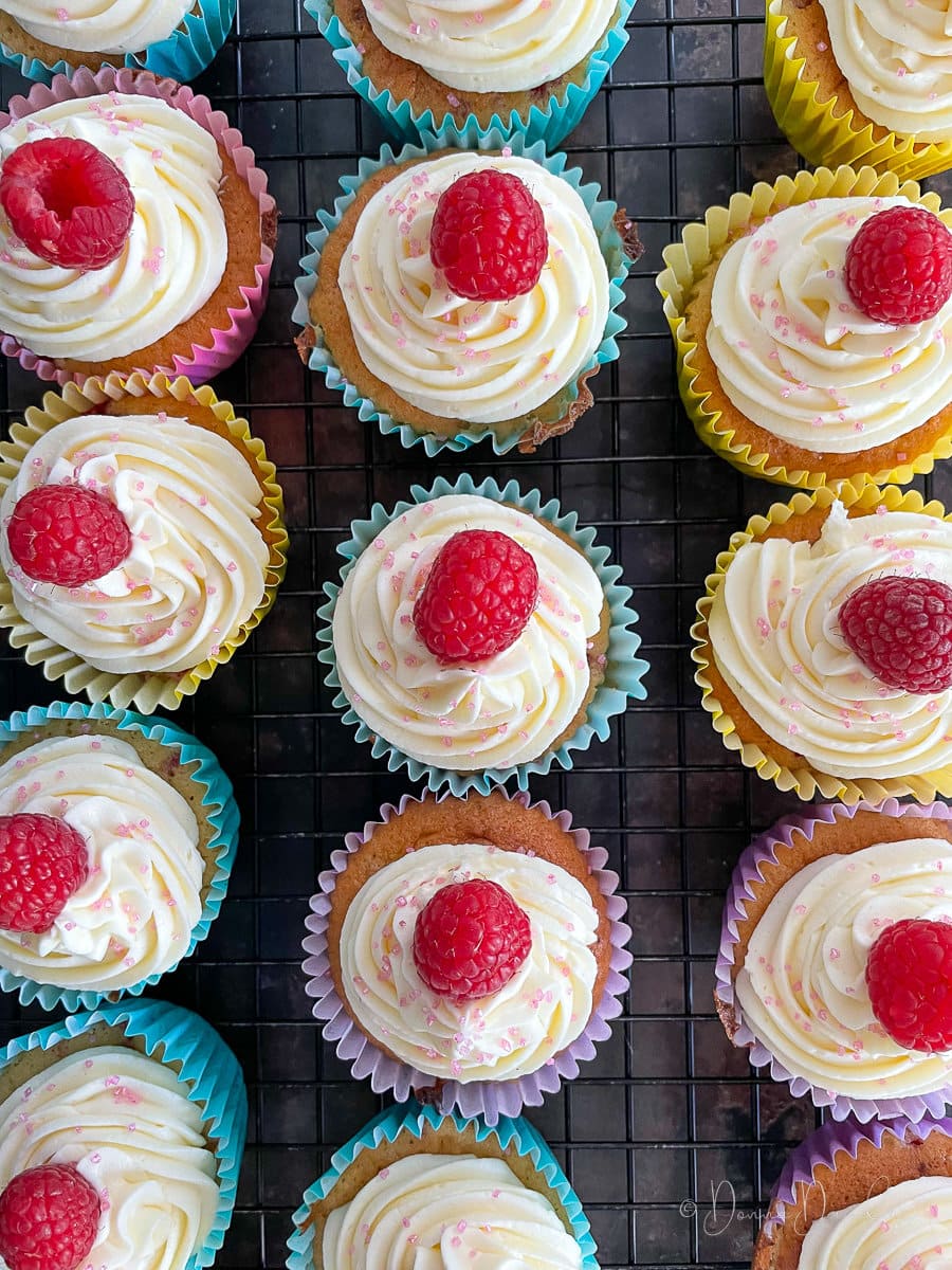 close up of cupcakes with icing and raspberries on top on wire rack.