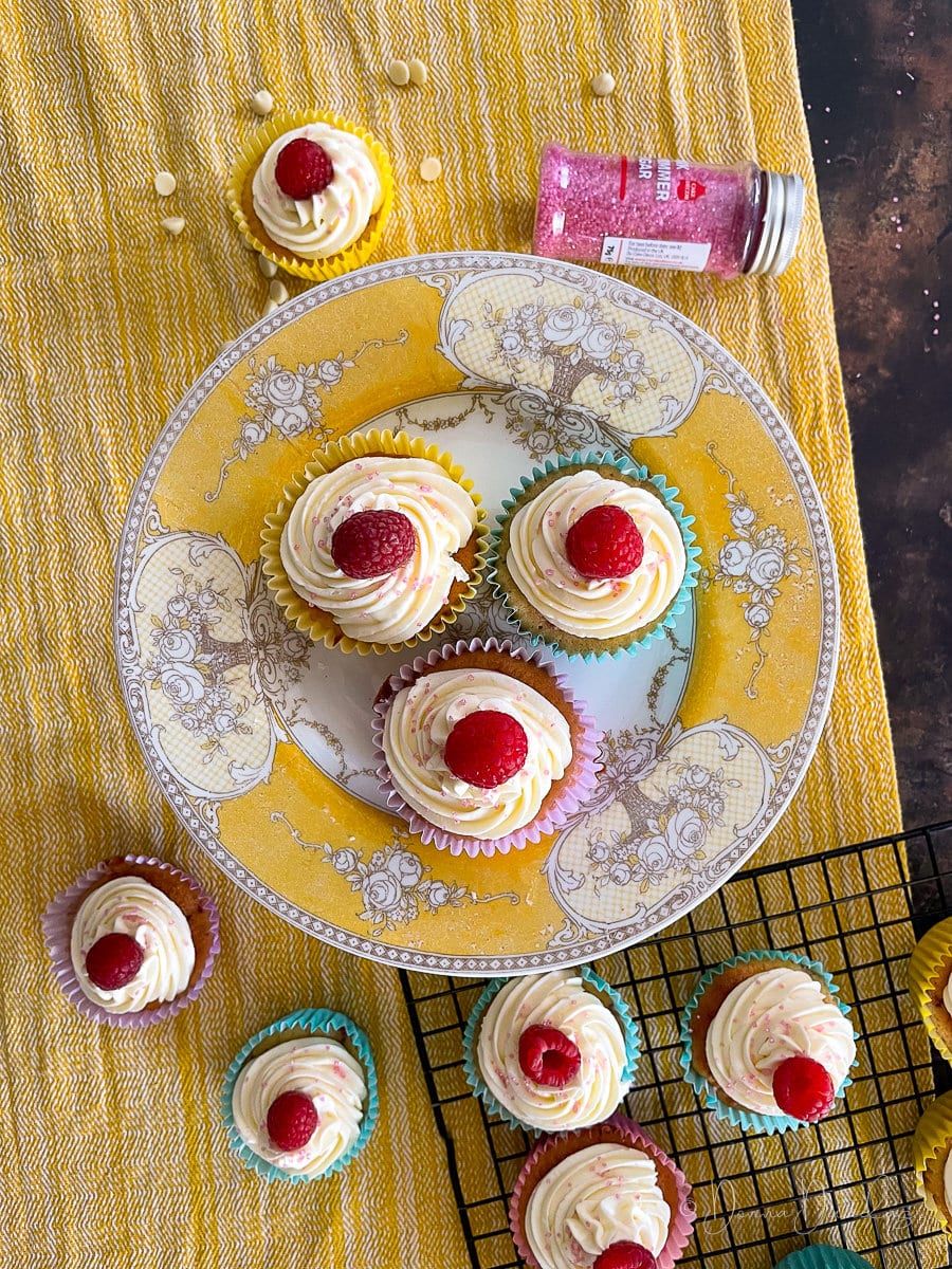 3 cupcakes on yellow printed cake stand. Other cakes, sprinkles and wire rack to the sides.