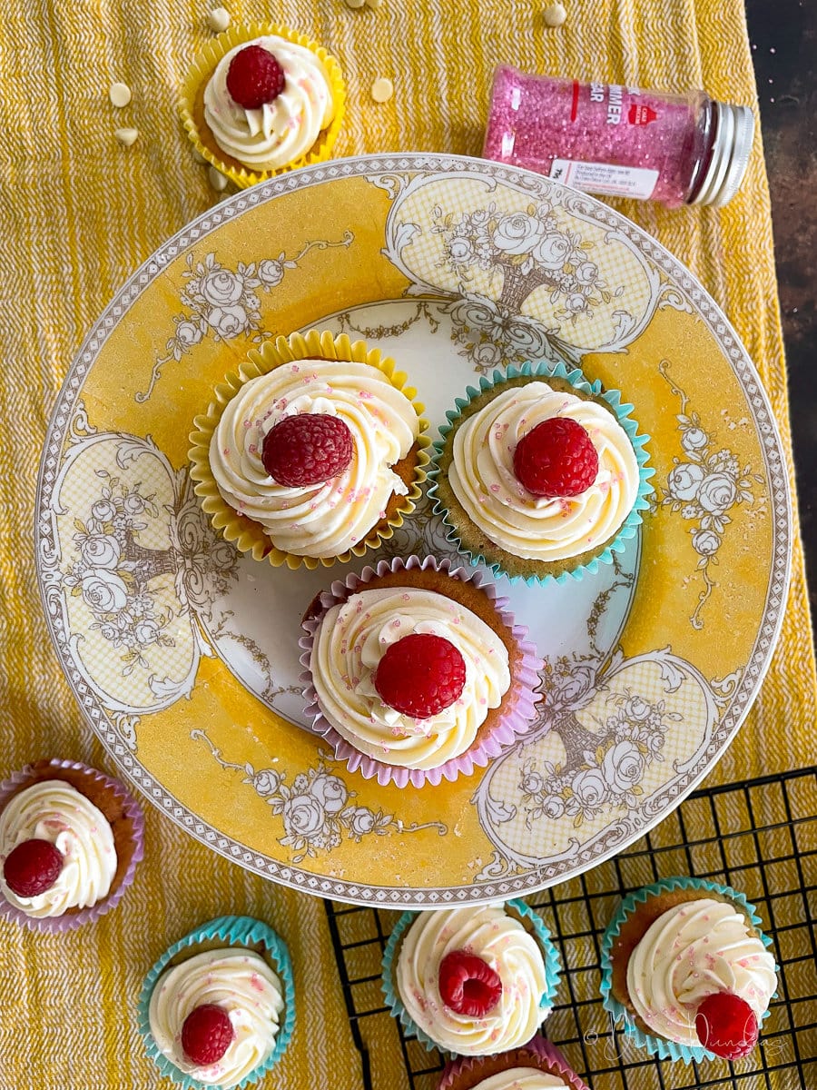 3 cupcakes on yellow printed cake stand. Other cakes, sprinkles and wire rack to the sides.