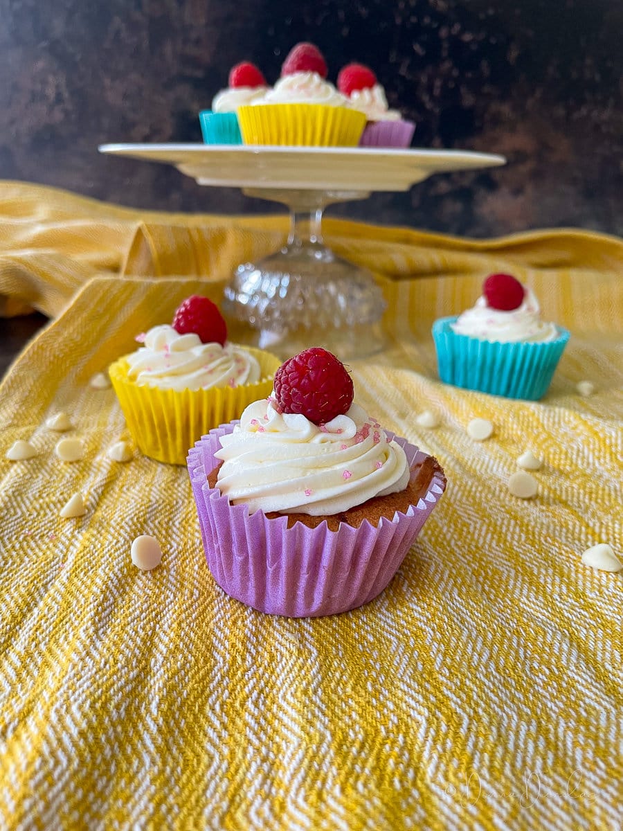 close up of cake topped with swirl of icing and raspberry. Other cakes and white choc chips in background.