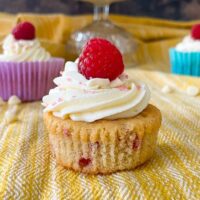 close up of cake topped with swirl of icing and raspberry, taken out of paper case. Other cakes and white choc chips in background.