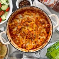 Spicy chicken pasta bake topped with cheese and baked in a cast iron dish. Bowls of salad and garlic bread to the sides. Mozzerella, salt and pepper, paprika and chilli also on table.