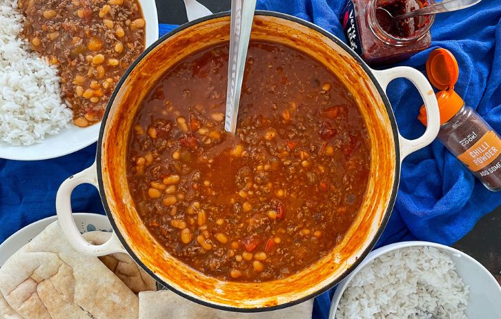 baked bean chilli in a cast iron dish with serving spoon. Served portion, bowl of rice, bread and chilli to the sides.