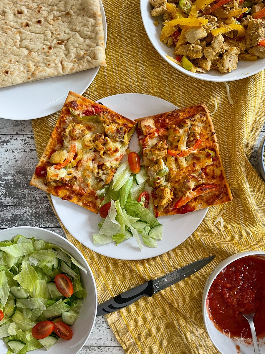 chicken tikka pizza cut in half on a plate with some salad. bread, salad, bowl of chicken and peppers and bowl of tomato sauce also on table.