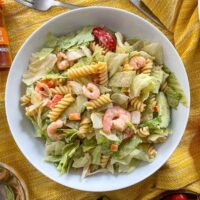 Prawn pasta salad in white pasta bowl. Pepper, dressing, hendersons relish and smoked paprika also on table.
