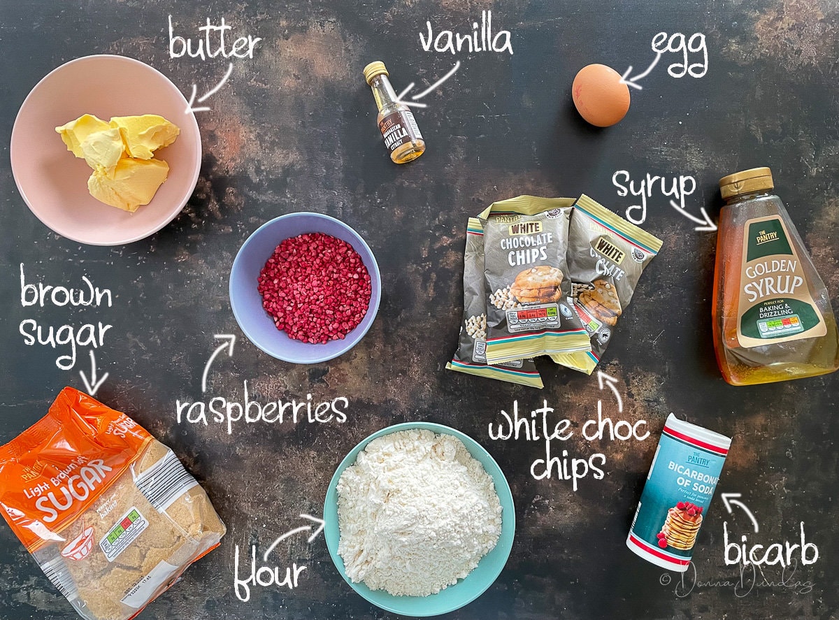 ingredients for white chocolate and raspberry cookies - butter, vanilla, egg, syrup, white chocolate chips, bicarb, flour, raspberries and brown sugar.