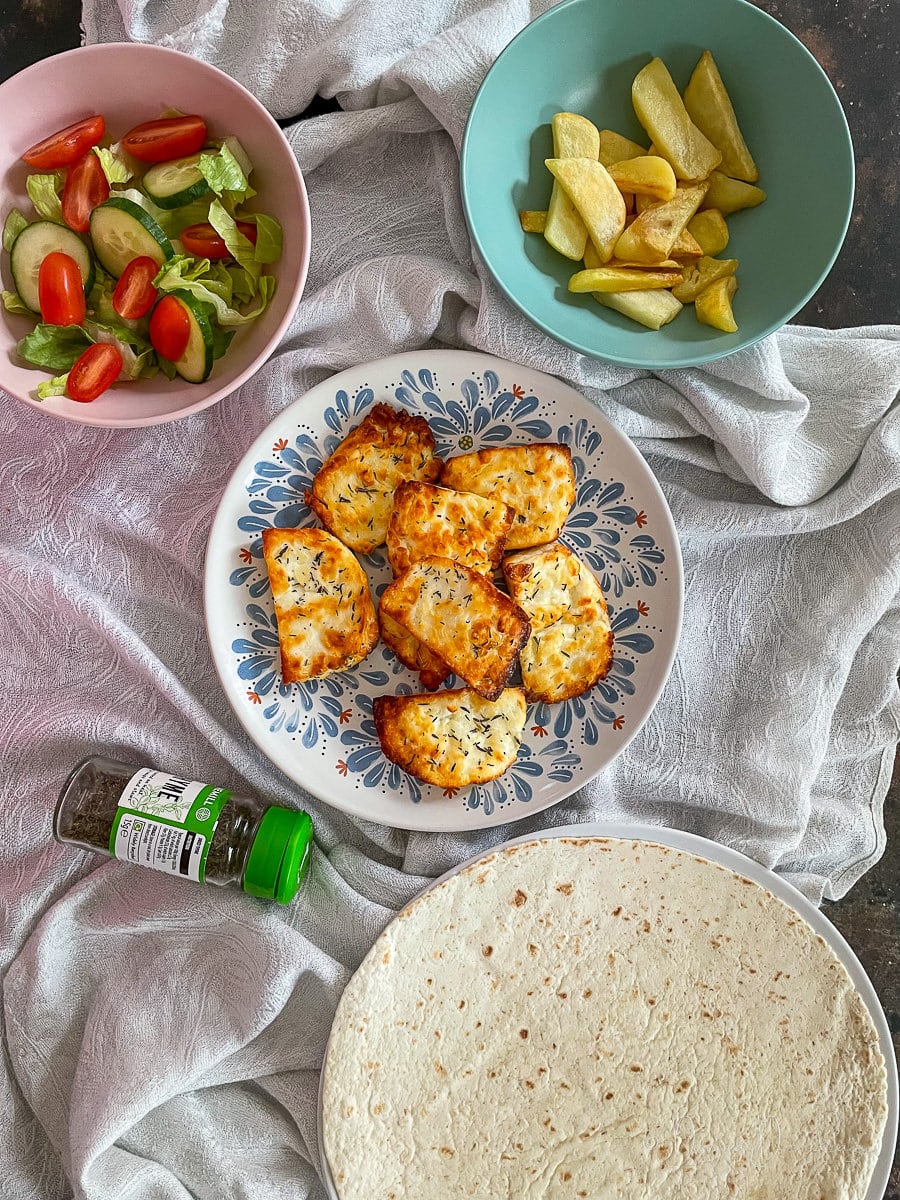 crispy halloumi slices on patterned plate. Salad, wraps and chips surrounding.