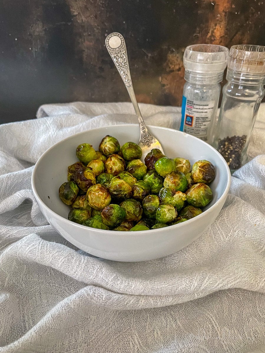 cooked sprouts in white bowl with fork. Salt and Pepper in the background.