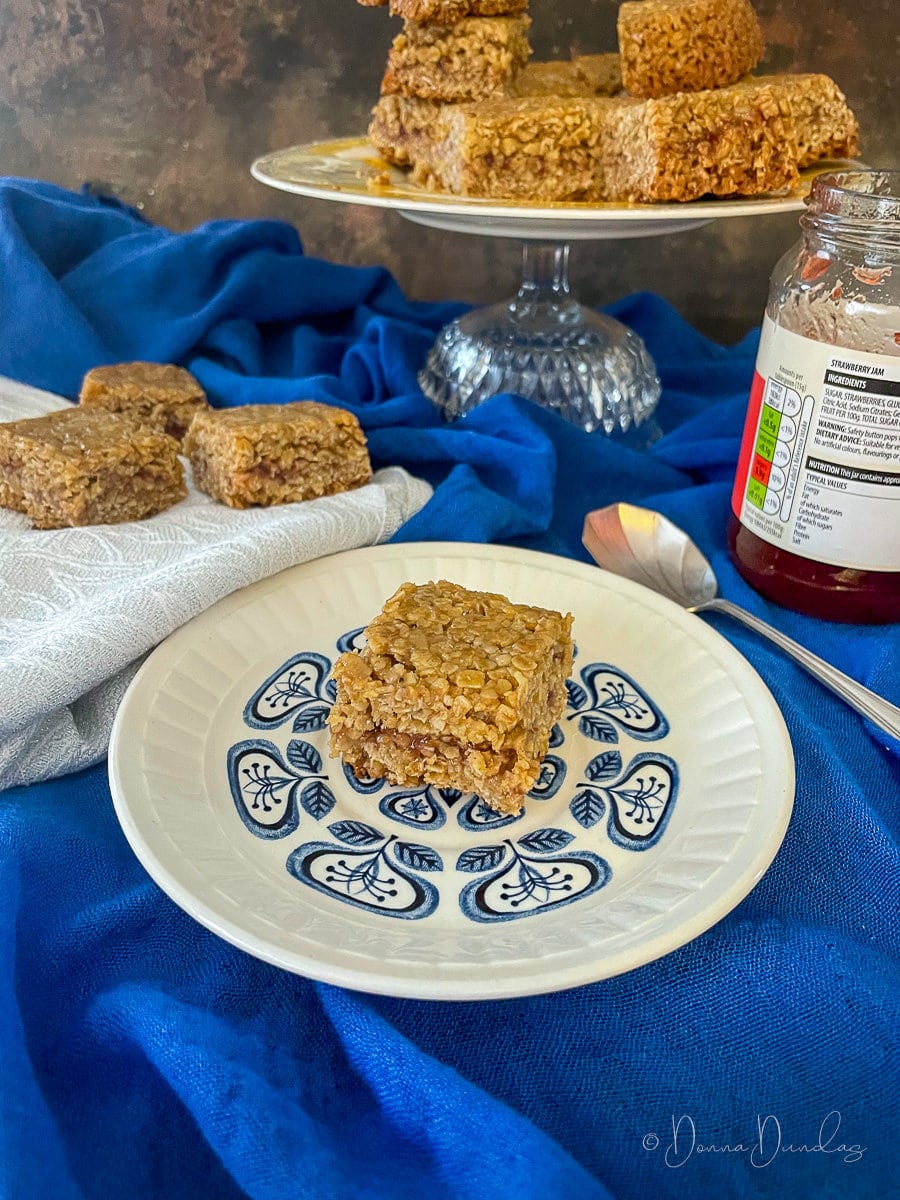 square of air fryer jam flapjack on plate, rest on cake stand in the background.