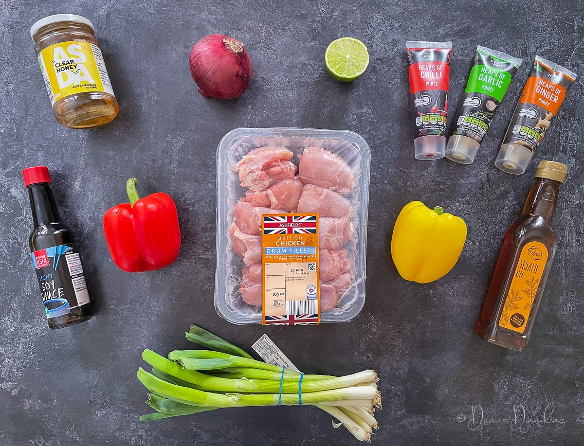 Ingredients for air fryer honey chilli chicken on table. Chicken fillets, spring onions, red and yellow peppers, soy sauce, sesame oil, garlic, ginger and chilli puree, lime, red onion and honey.