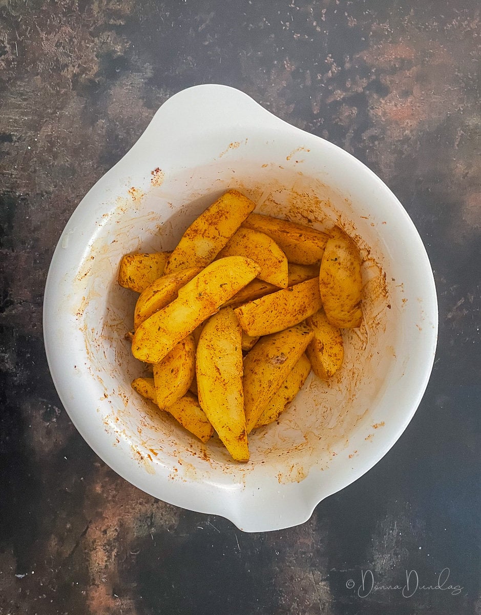 uncooked potato wedges seasoned in white mixing bowl.