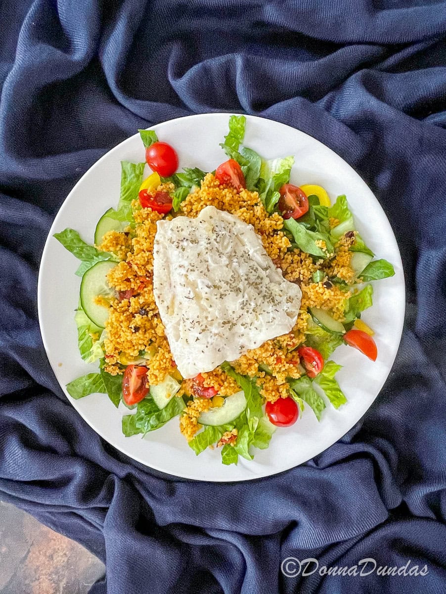Top view of white plate with lettuce, tomato, cucumber, cous cous and a cod fillet.