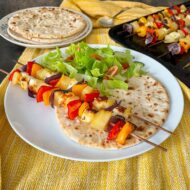 2 skewers wtih halloumi cheese, pineapple chunks, peppers and onions on a plate with a flatbread and salad.