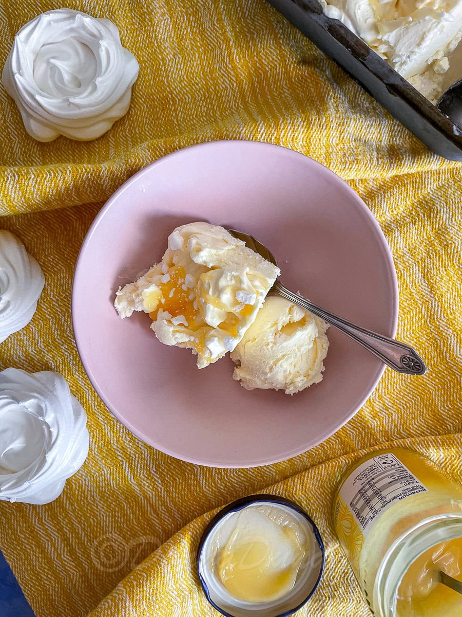 Ice cream in bowl topped with lemon curd and meringue. Rest of ice cream in loaf tin in the background. Lemon curd and merignue nests also on table.