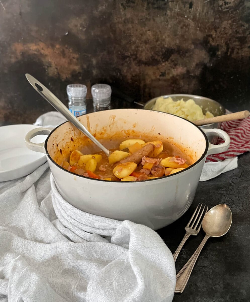 Sausage and apple casserole with cider in a cooking dish with a serving spoon, pan of mashed potato in the background.